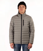 A man wearing a taupe quilted puffer jacket with a high collar, black zipper details, a black beanie, and blue jeans, standing against a plain white background.