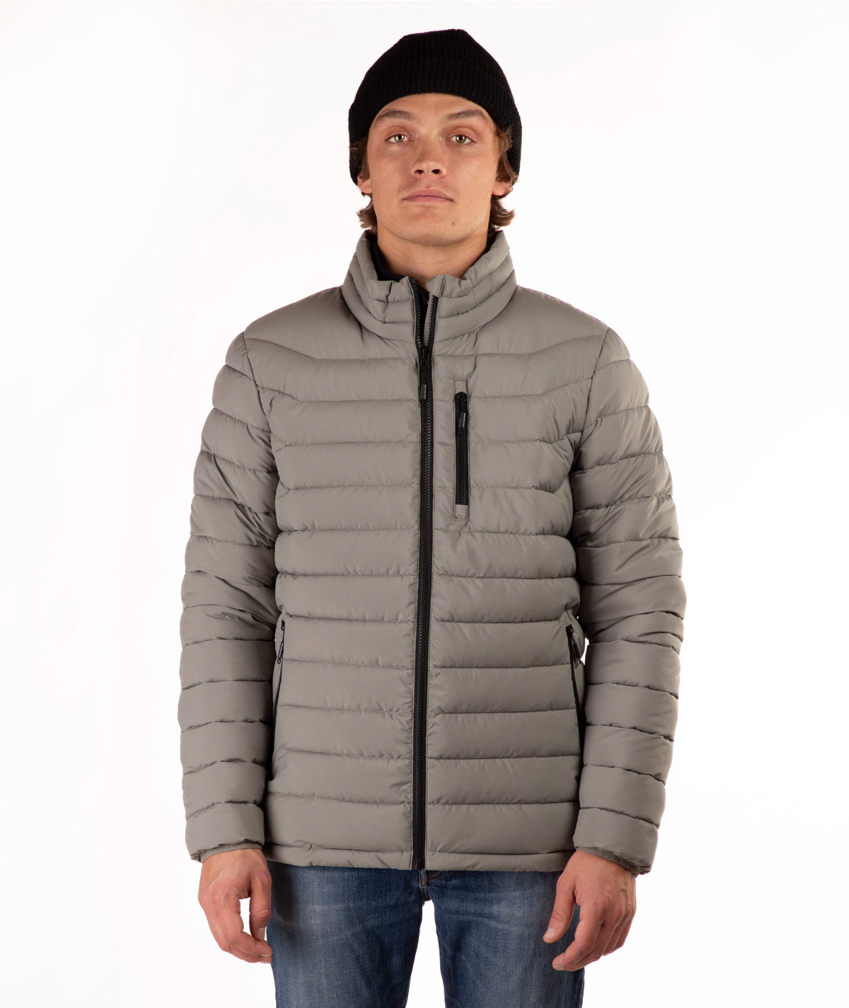 A man wearing a taupe quilted puffer jacket with a high collar, black zipper details, a black beanie, and blue jeans, standing against a plain white background.