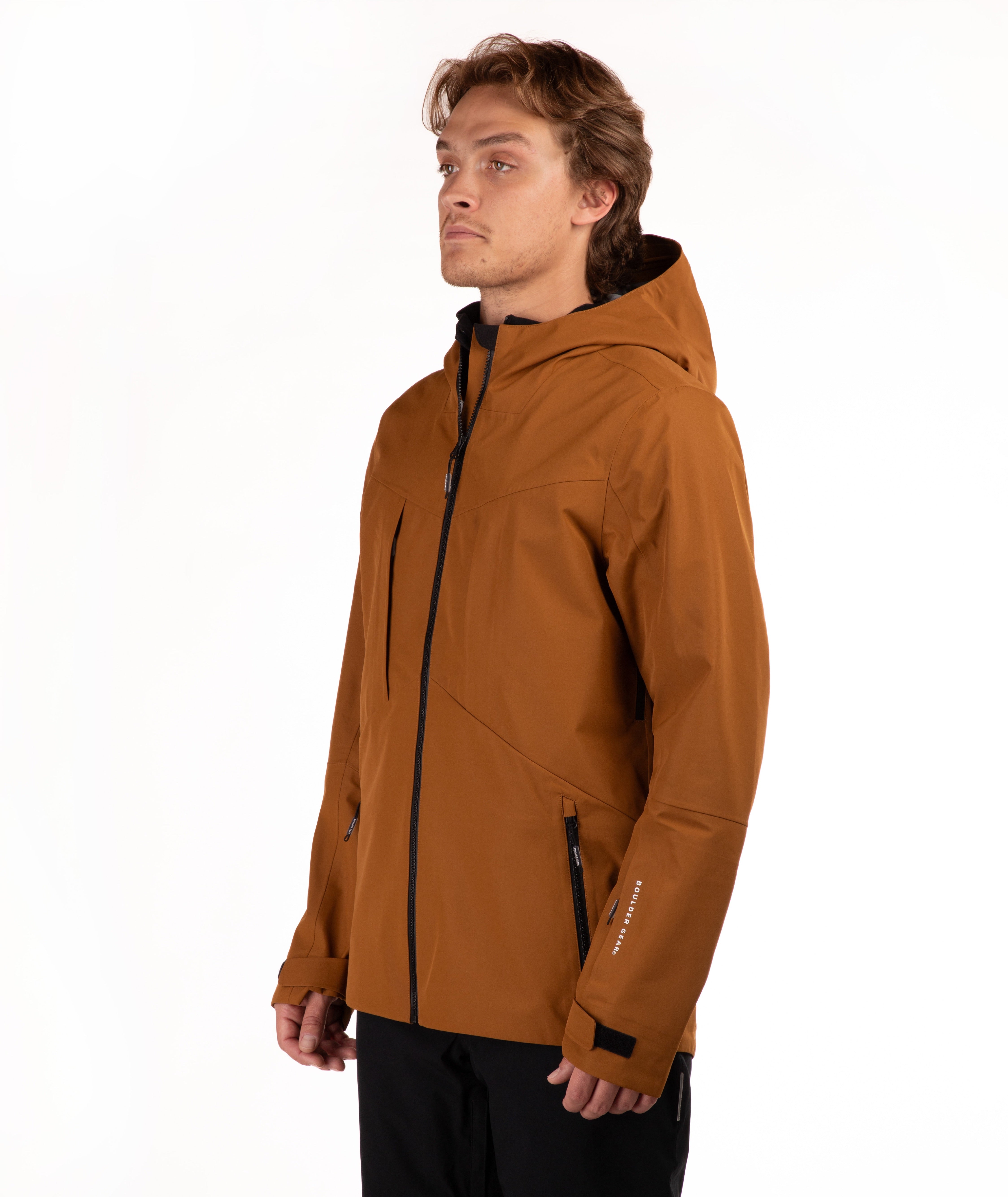A man wearing a brown hooded technical jacket with black zippers and black pants, standing against a plain white studio background.