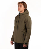 A man wearing an olive green technical outdoor jacket with black zippers and black pants, standing against a plain white studio background.