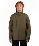 A man wearing an olive green technical outdoor jacket with black zippers and black pants, standing against a plain white studio background.
