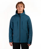 A man wearing a teal blue hooded ski jacket with black zippers, standing against a plain white studio background.