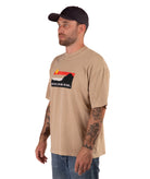 Man wearing a beige t-shirt with a logo and text, standing against a white background