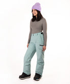 A woman modeling snow pants in a studio setting, wearing a gray long-sleeve top, a light purple knit beanie, and black snow boots against a plain white background.
