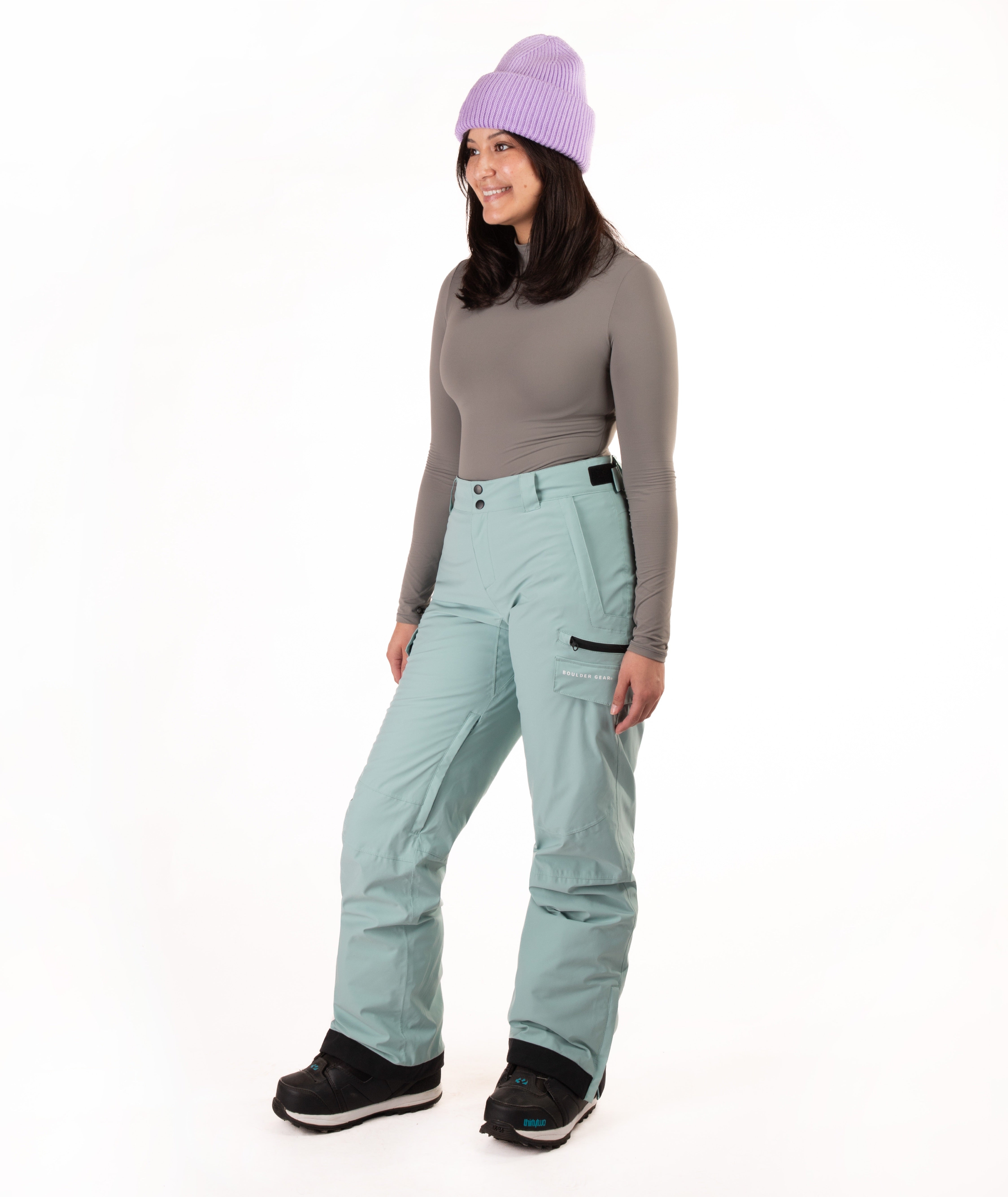 A woman modeling snow pants in a studio setting, wearing a gray long-sleeve top, a light purple knit beanie, and black snow boots against a plain white background.