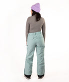 A model shown from the back wearing light sage green snow pants, a gray long-sleeve top, a lavender knit beanie, and black boots, photographed against a seamless white studio background.