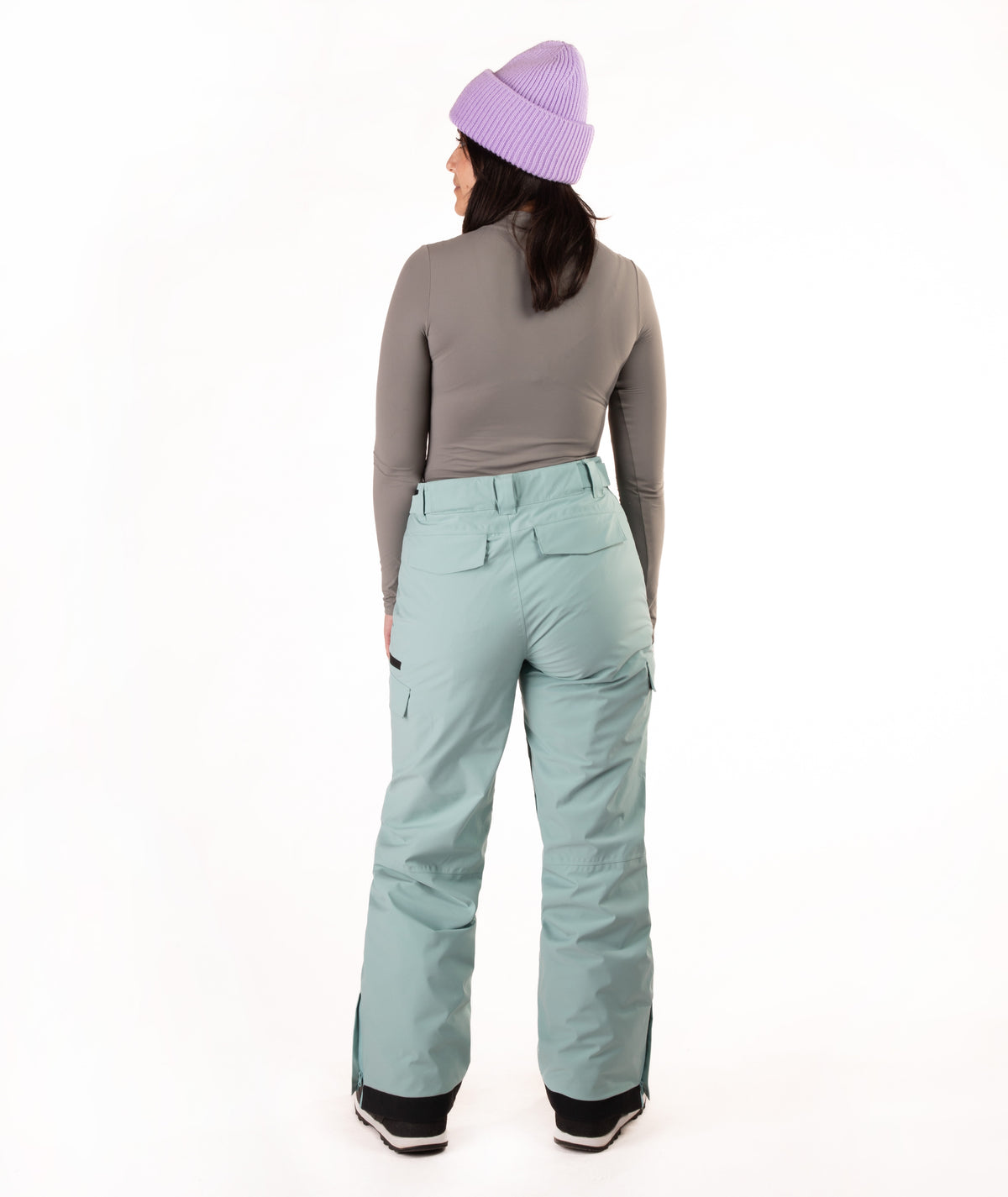 A model shown from the back wearing light sage green snow pants, a gray long-sleeve top, a lavender knit beanie, and black boots, photographed against a seamless white studio background.