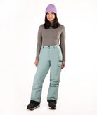 A woman modeling light sage green snow pants with a gray long-sleeve top, a purple knit beanie, and black snow boots, standing against a solid white background.