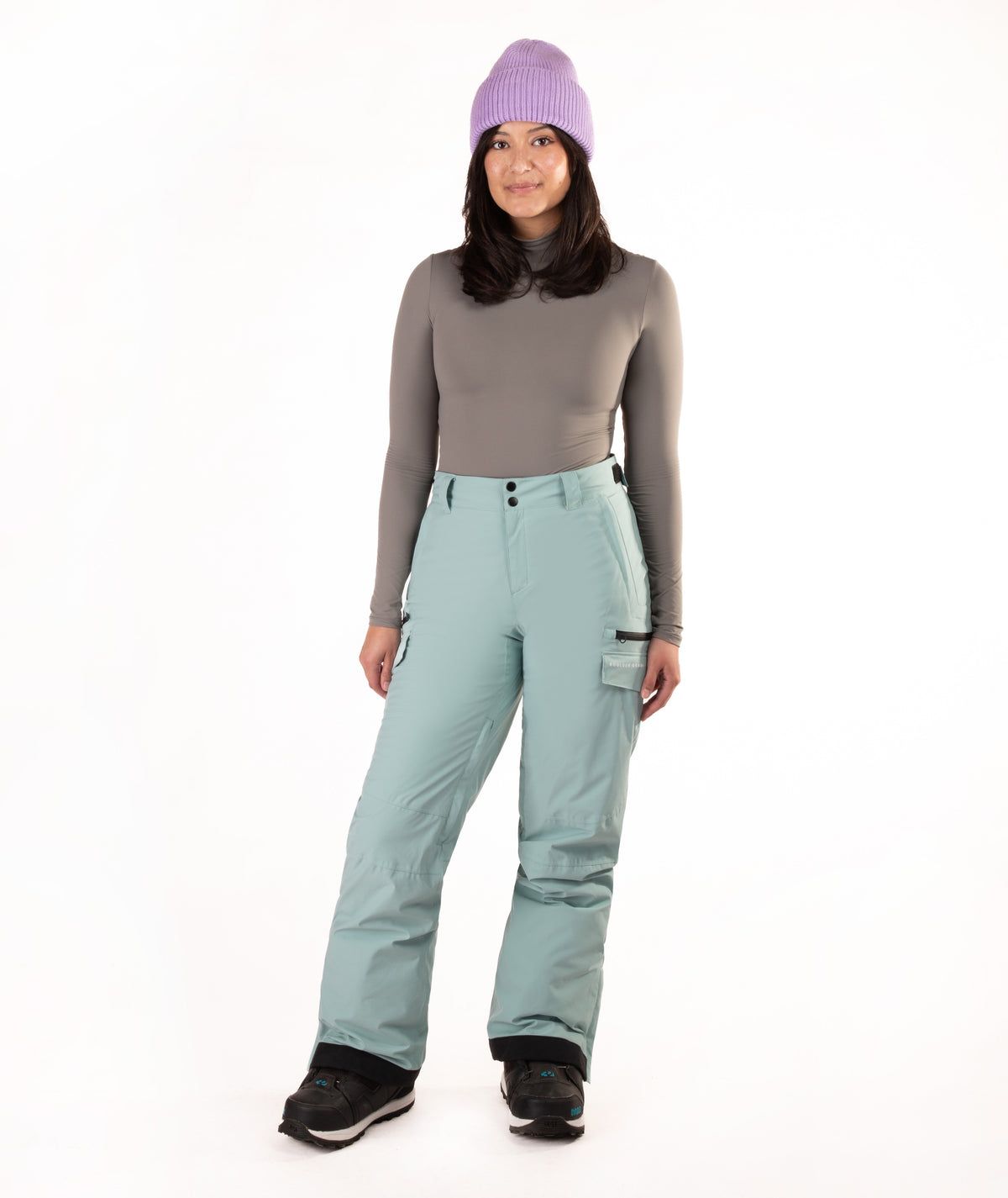 A woman modeling light sage green snow pants with a gray long-sleeve top, a purple knit beanie, and black snow boots, standing against a solid white background.