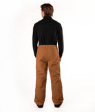 A man viewed from behind wearing a black long-sleeve shirt and brown snow pants against a white studio background.