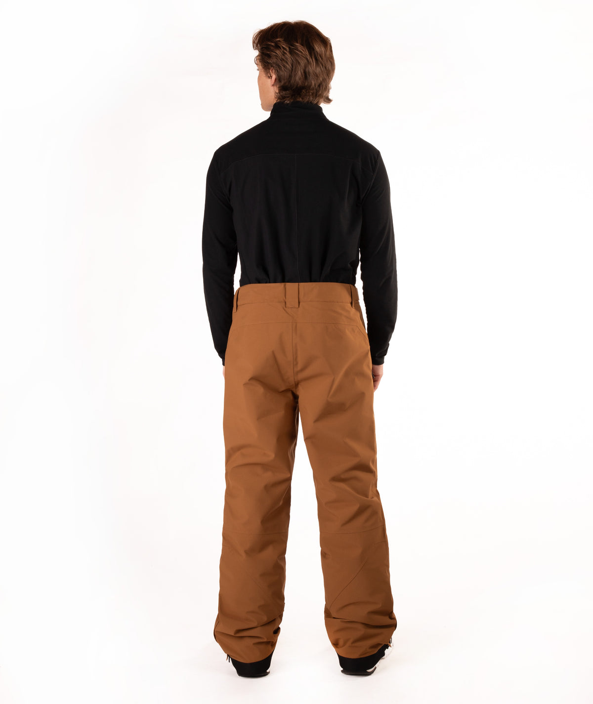 A man viewed from behind wearing a black long-sleeve shirt and brown snow pants against a white studio background.