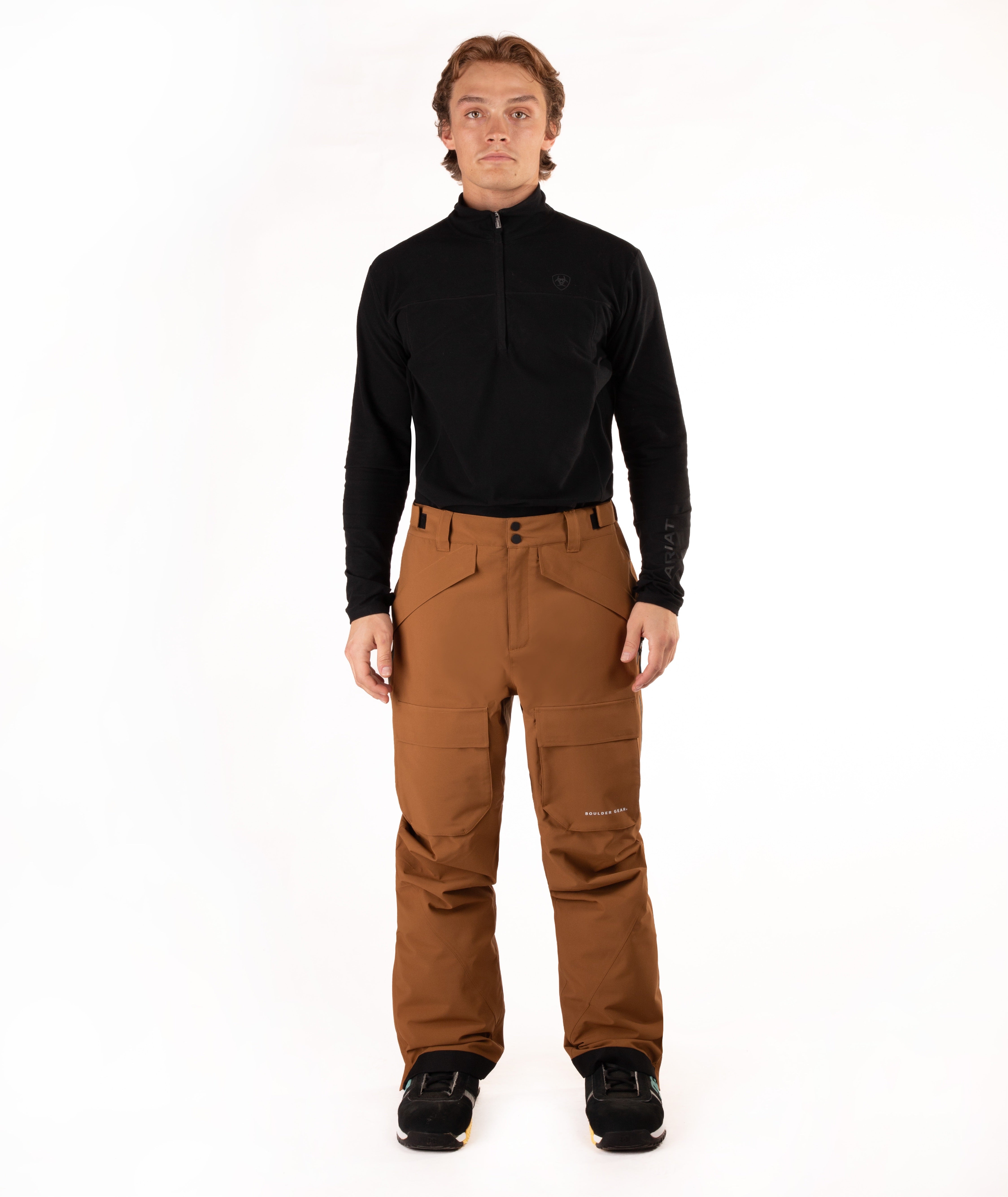 A man modeling a black long-sleeve quarter-zip top and brown snow pants with black snow shoes, standing against a solid white studio background.