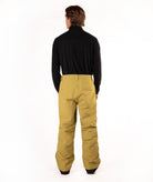 A man viewed from behind wearing a black long-sleeve shirt and mustard yellow snow pants against a white studio background.