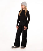 A young girl modeling a black long-sleeve top, black snow pants, a light gray beanie, and gray boots against a plain white studio background.