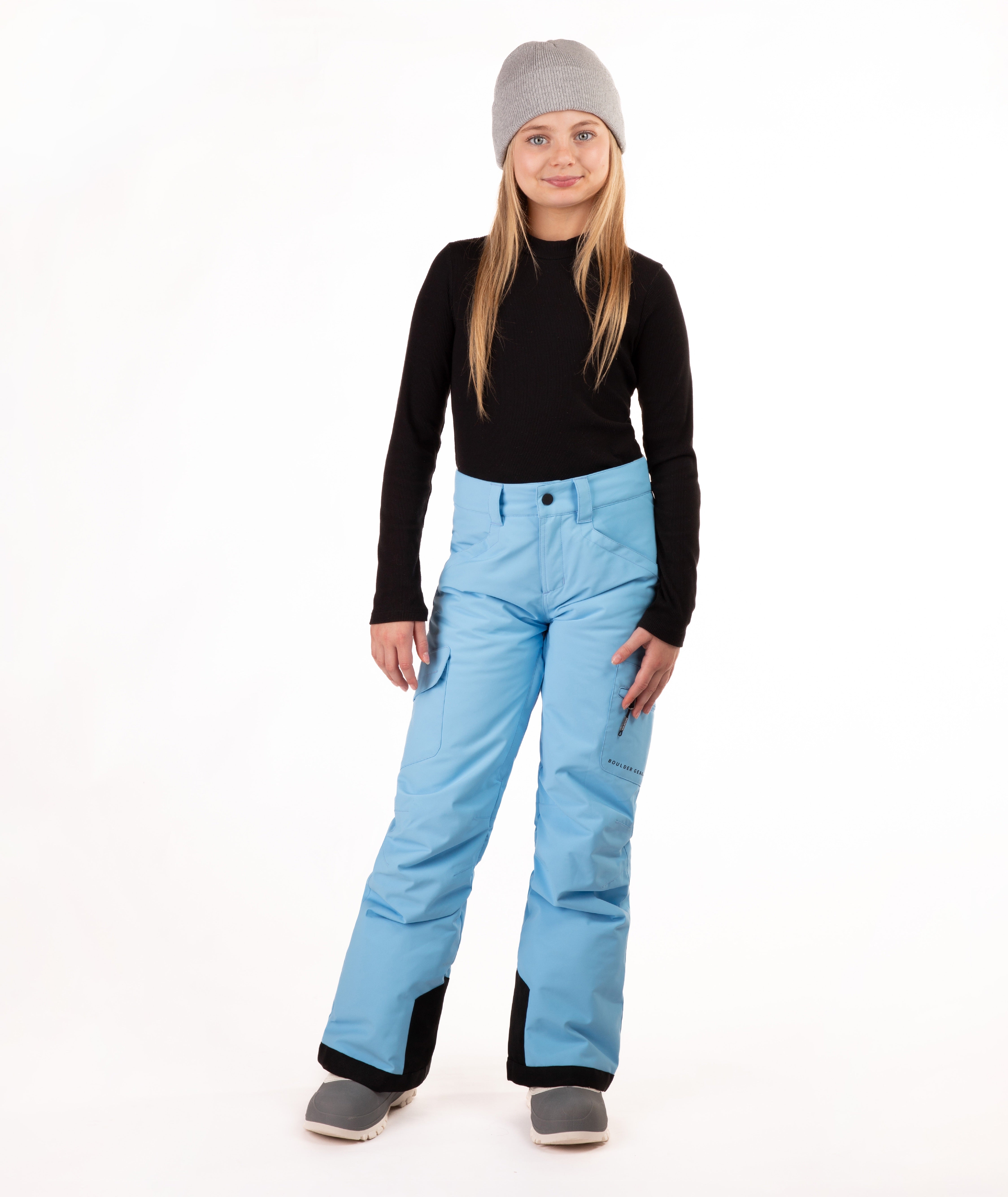 A young girl modeling a pair of light blue ski pants, a black long-sleeve top, and a gray beanie against a white studio background.