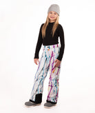 A young girl modeling snow pants with a colorful paint-splatter pattern, paired with a black long-sleeve top, gray beanie, and gray boots, standing against a white studio background.