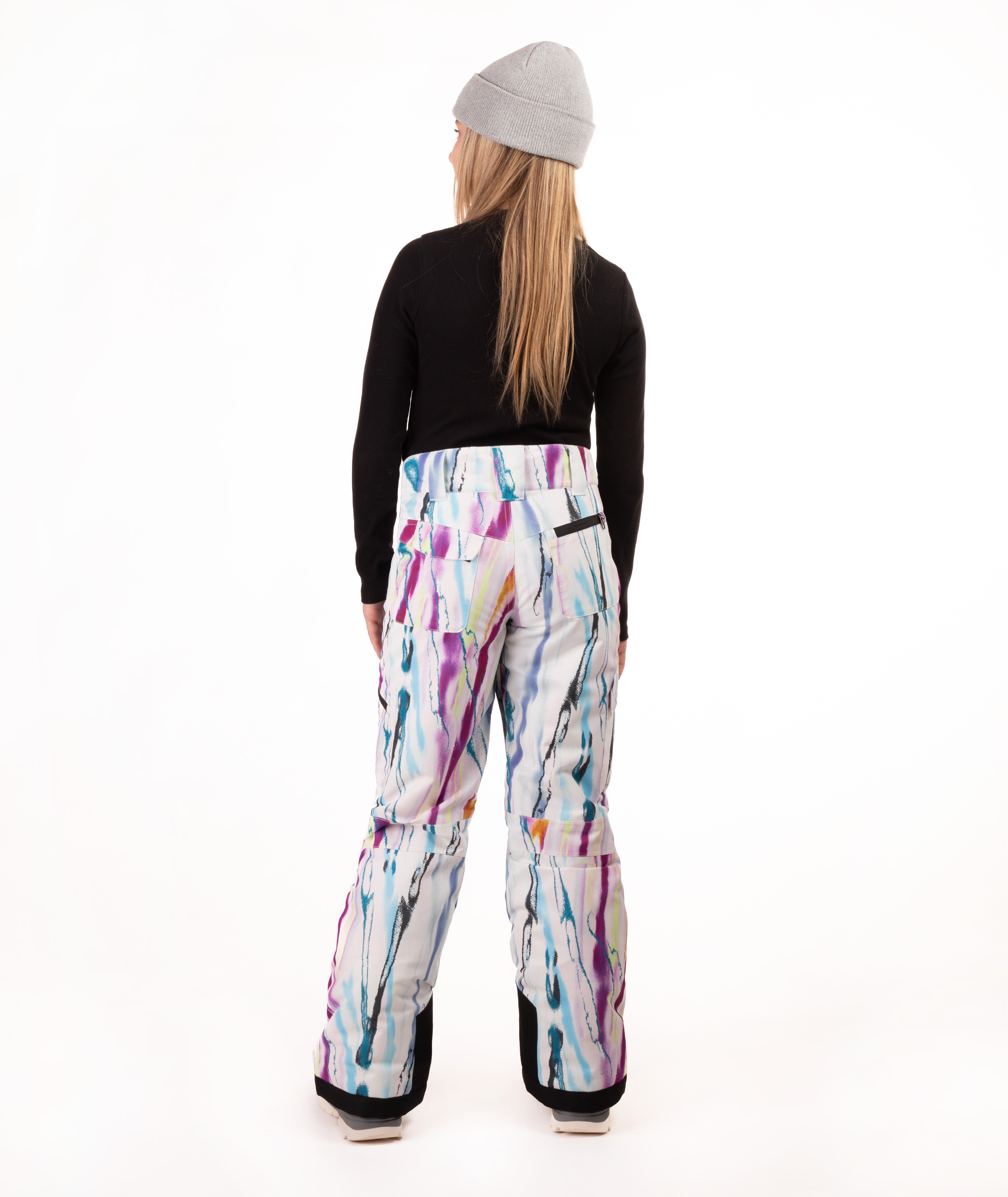 A model shown from the back wearing  snow pants with a colorful paint-splatter pattern, a black long-sleeve top, and a gray beanie against a white studio background.