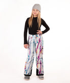 A young girl modeling snow pants with a colorful paint-splatter pattern, paired with a black long-sleeve top, gray beanie, and gray boots, standing against a white studio background.