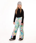 A young girl modeling colorful tie-dye snow pants, a black long-sleeve shirt, and a gray beanie against a white studio background.