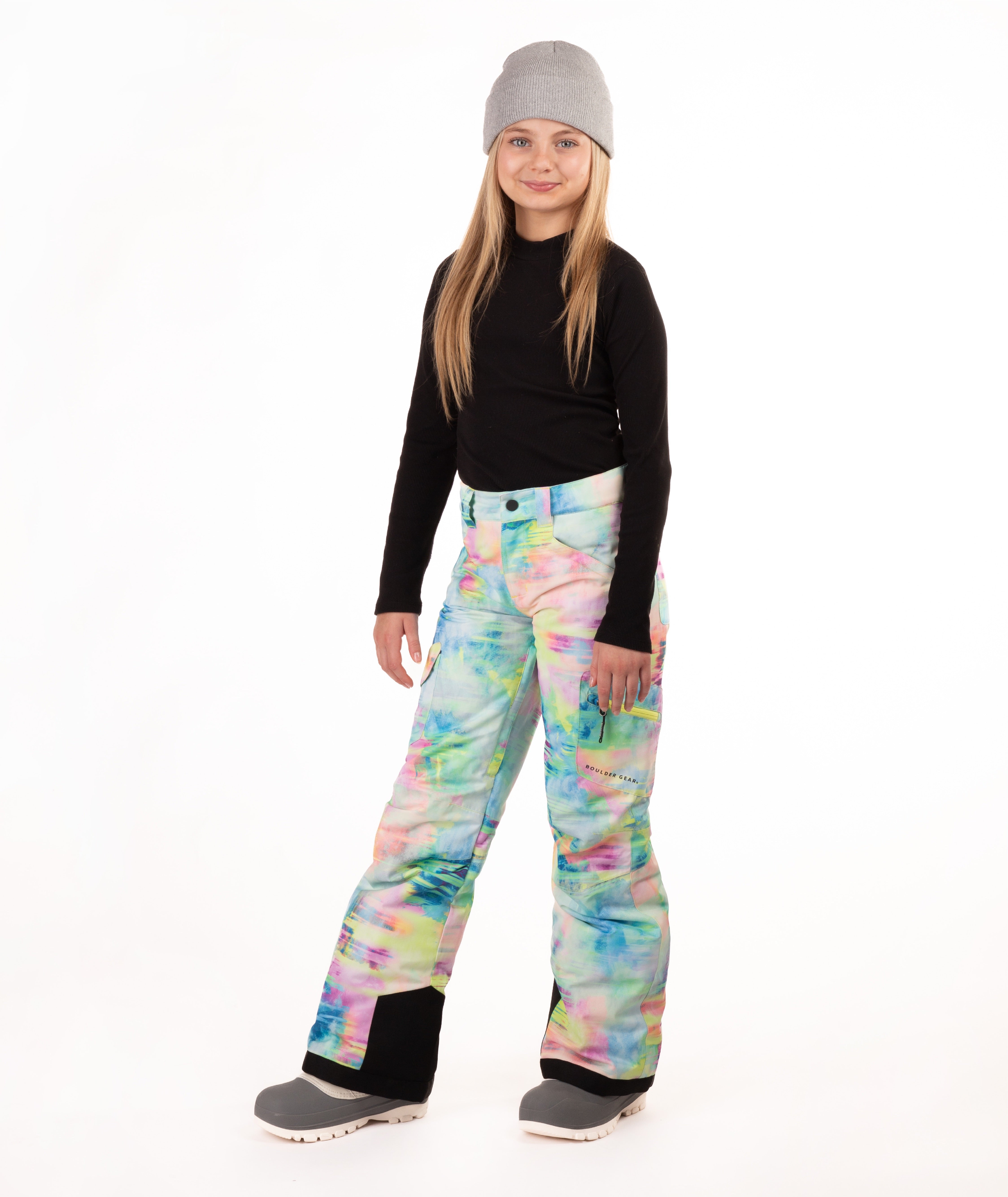 A young girl modeling colorful tie-dye snow pants, a black long-sleeve shirt, and a gray beanie against a white studio background.