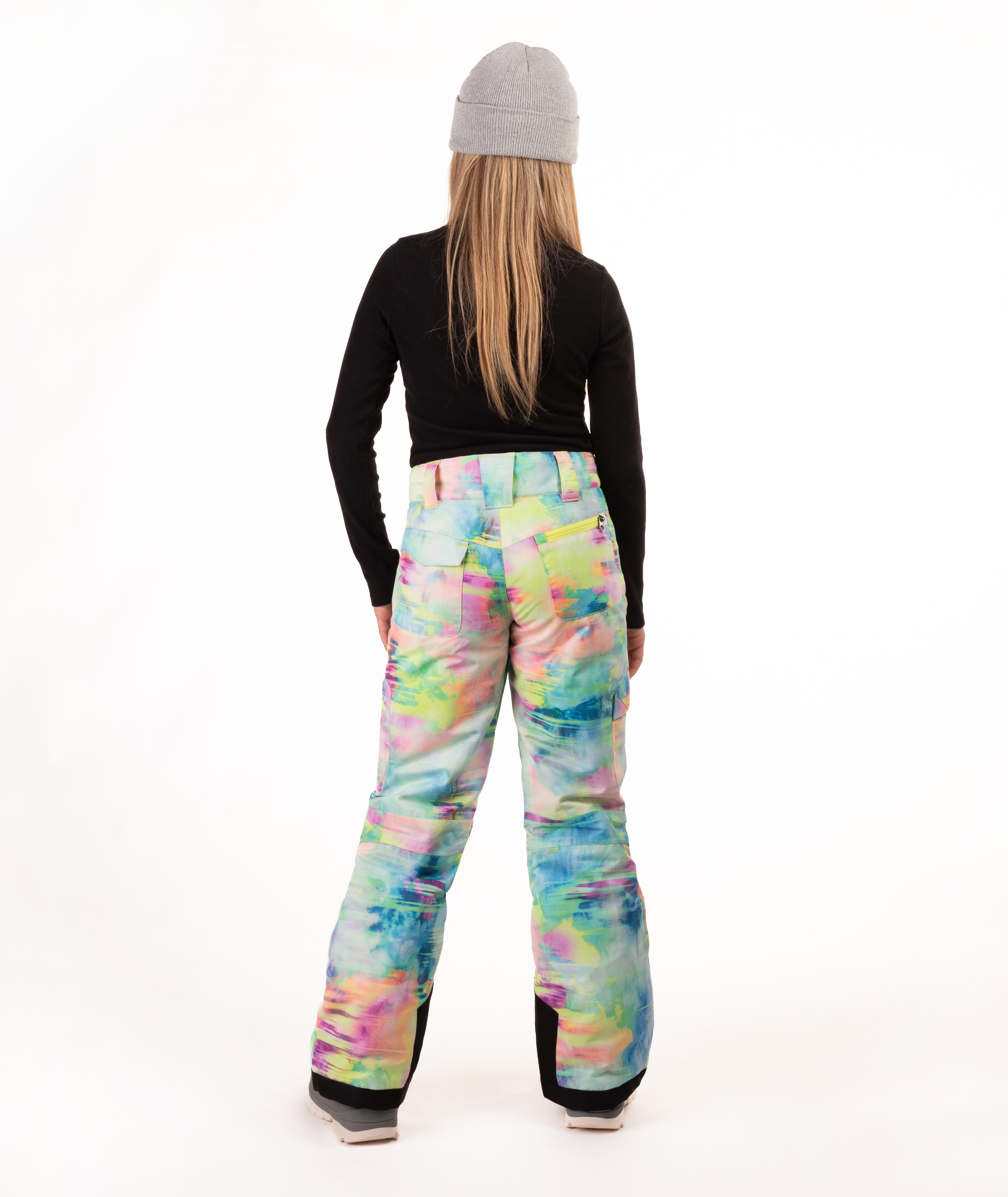 A person shown from the back wearing colorful snow pants with a pastel tie-dye pattern, a black long-sleeve top, and a gray beanie against a plain white studio background.