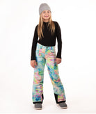 A young girl modeling colorful tie-dye snow pants, a black long-sleeve shirt, and a gray beanie against a white studio background.