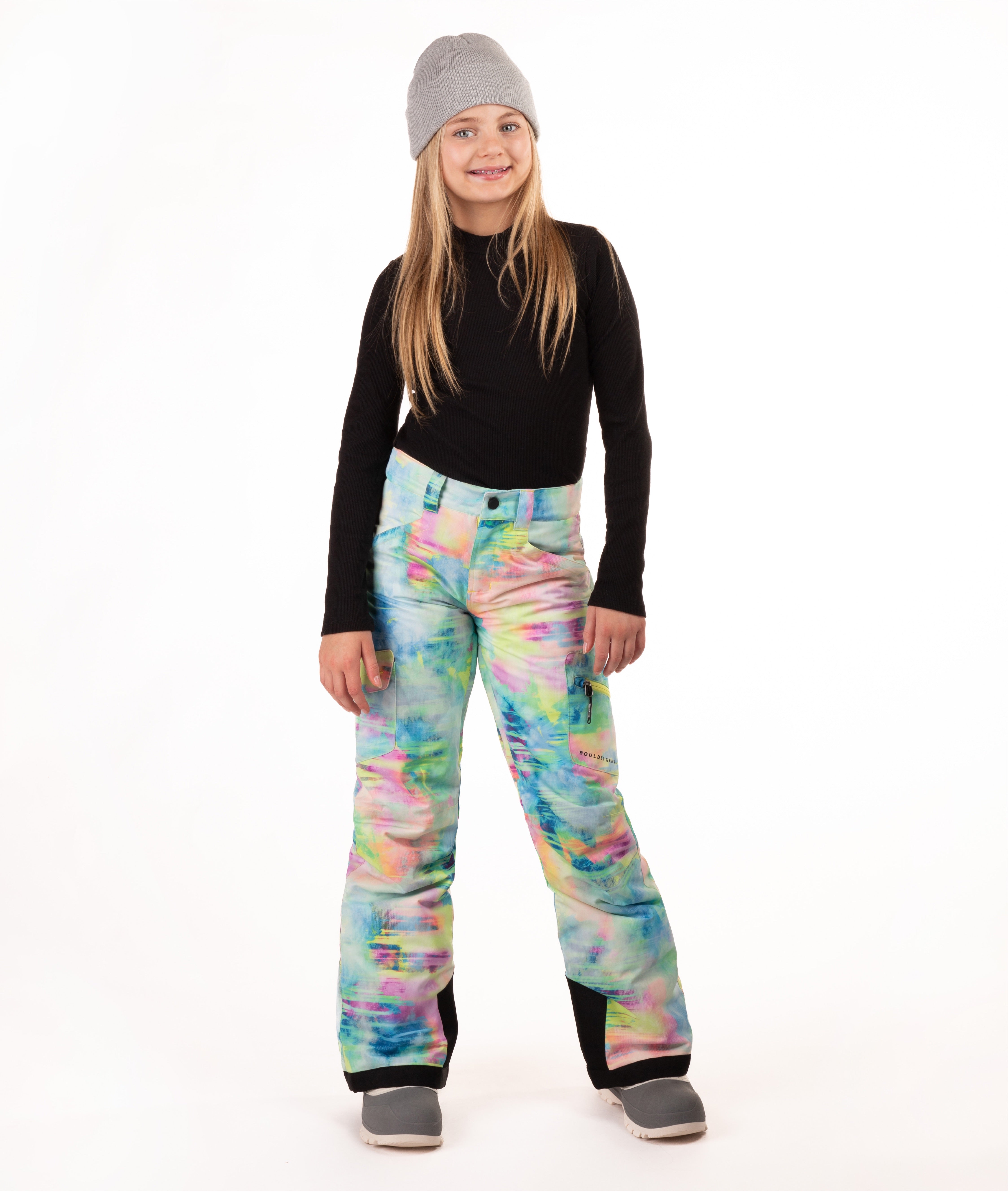 A young girl modeling colorful tie-dye snow pants, a black long-sleeve shirt, and a gray beanie against a white studio background.