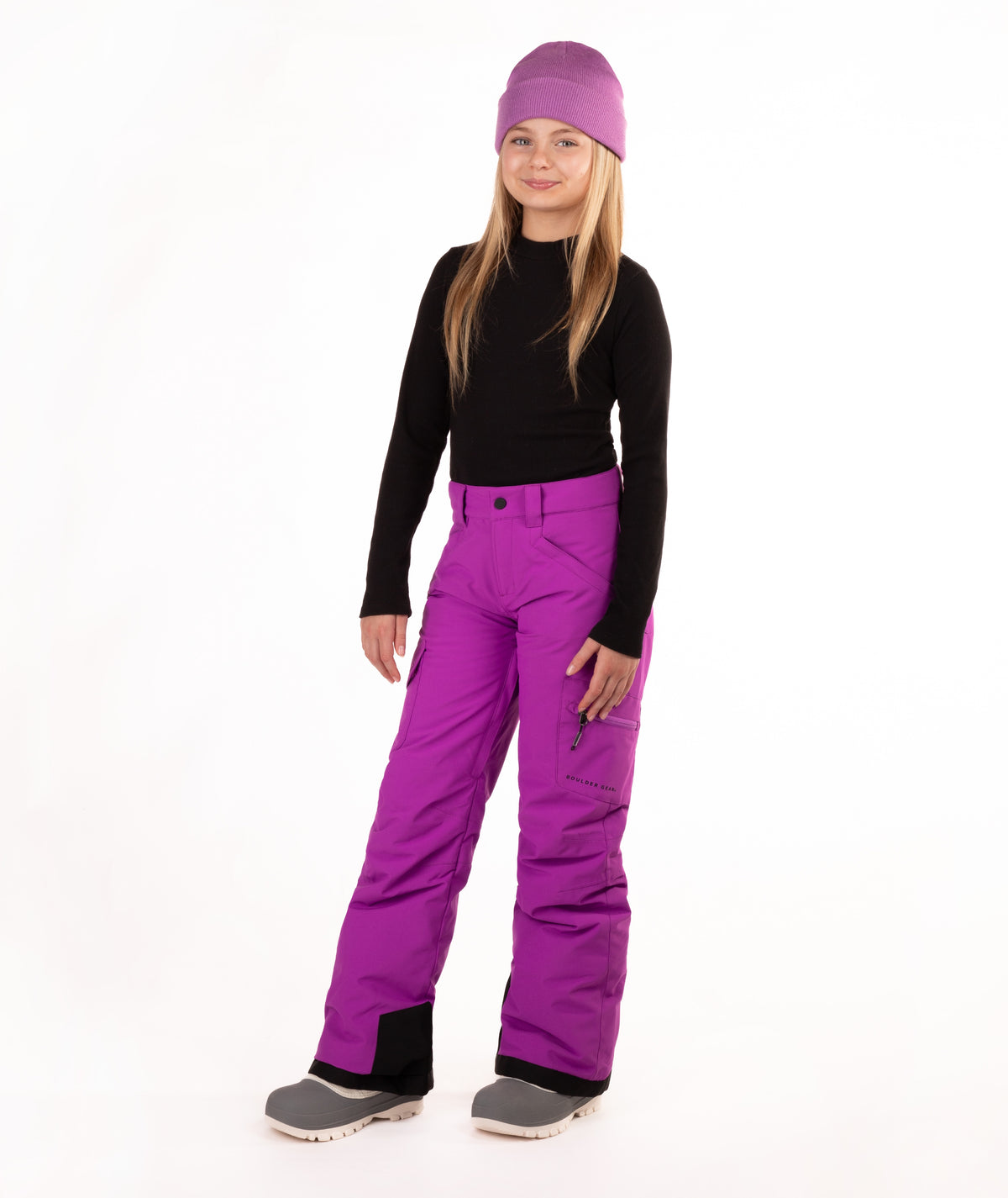 A young girl standing against a white studio background, wearing a purple knit beanie, a black long-sleeve top, bright purple snow pants, and gray boots.