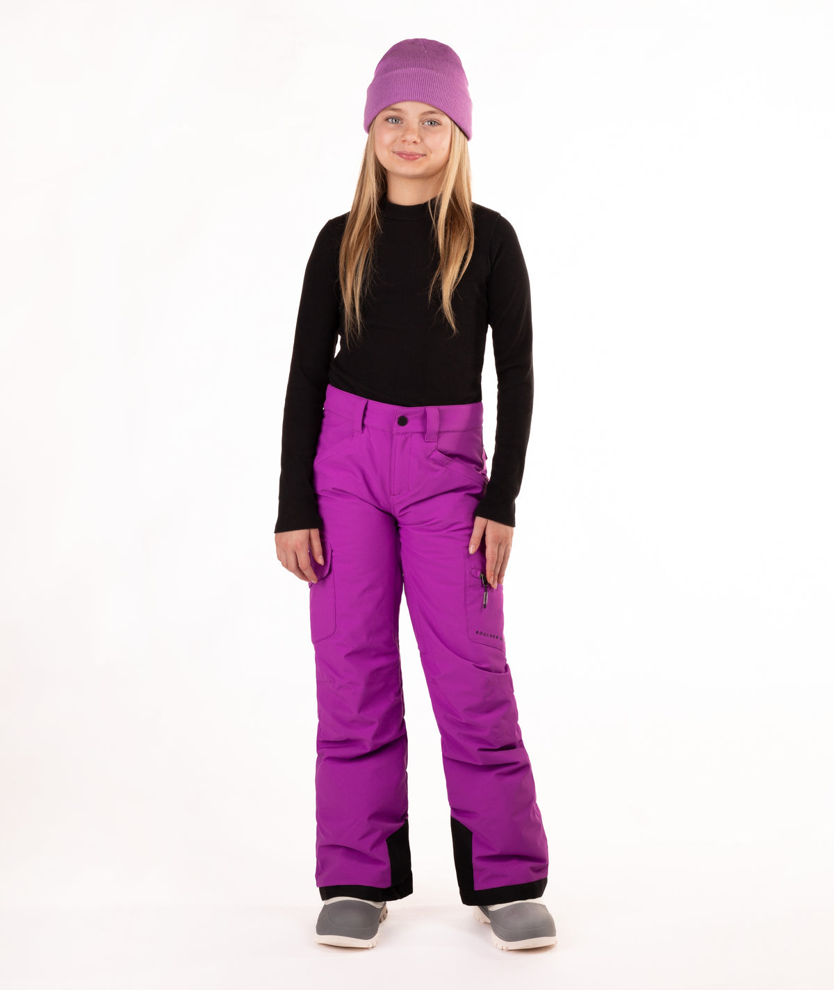 A young girl standing against a white studio background, wearing a purple knit beanie, a black long-sleeve top, bright purple snow pants, and gray boots.