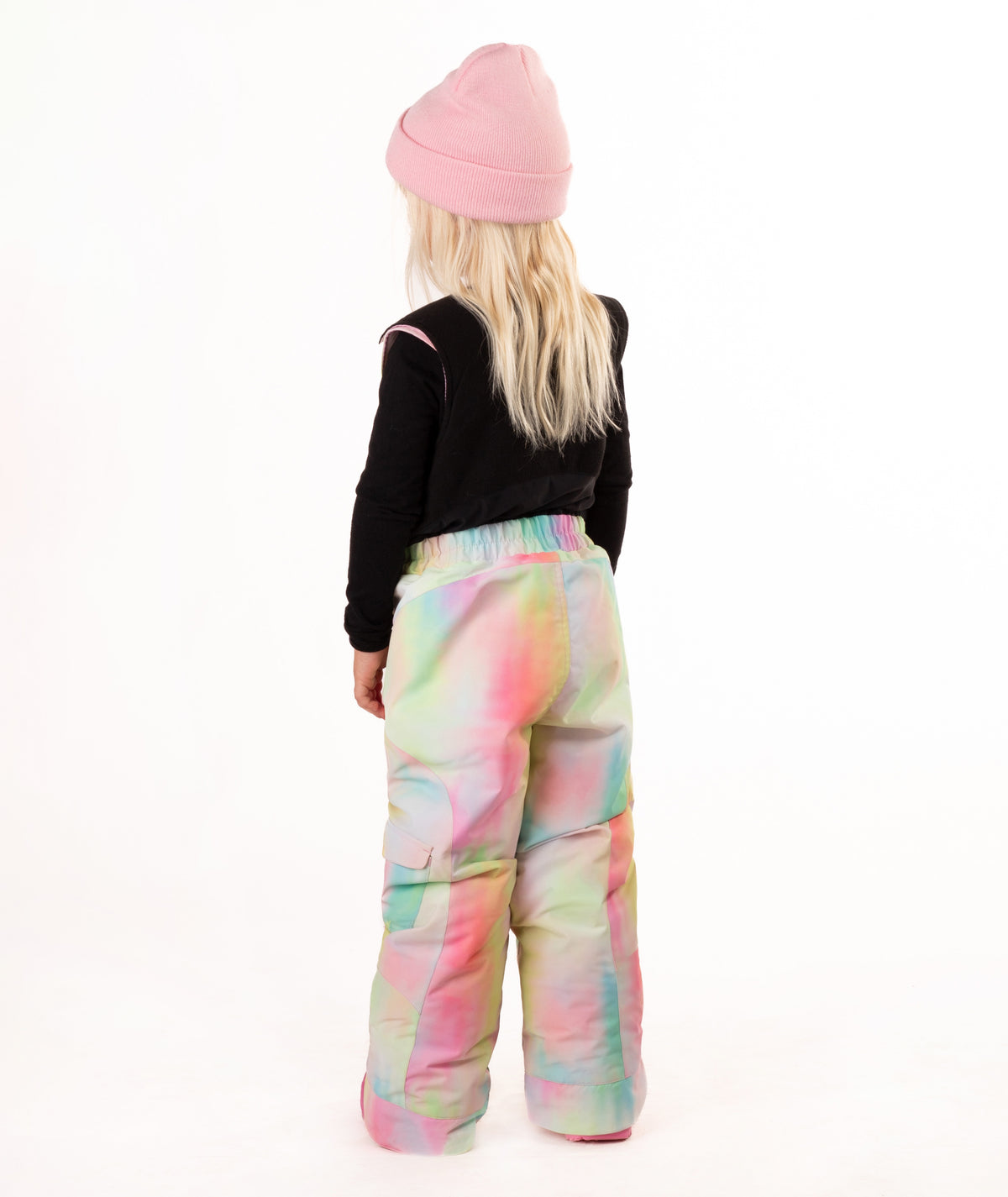 A child shown from the back wearing a pink beanie, a black long-sleeve top, and snow bib with a pastel tie-dye pattern in pink, green, blue, and yellow, against a plain white background.