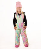 A young girl wearing a pastel rainbow tie-dye snow bib over a black long-sleeve shirt, a pink beanie, and pink boots, standing against a white studio background.