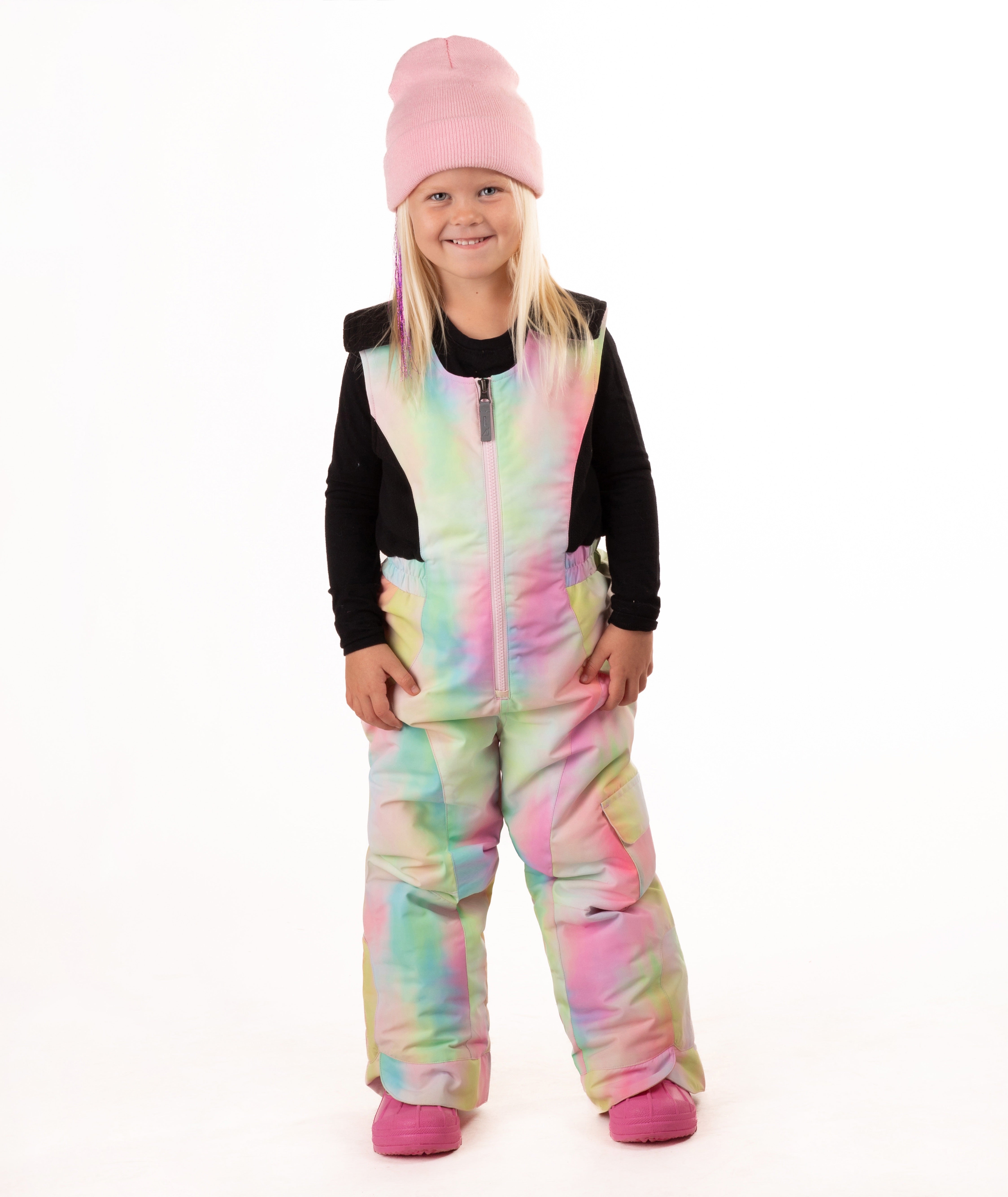 A young girl wearing a pastel rainbow tie-dye snow bib over a black long-sleeve shirt, a pink beanie, and pink boots, standing against a white studio background.