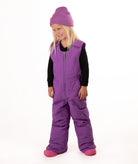 A young child wearing a purple snow bib over a black long-sleeve shirt, a matching purple knit beanie, and pink boots, standing against a plain white studio background.