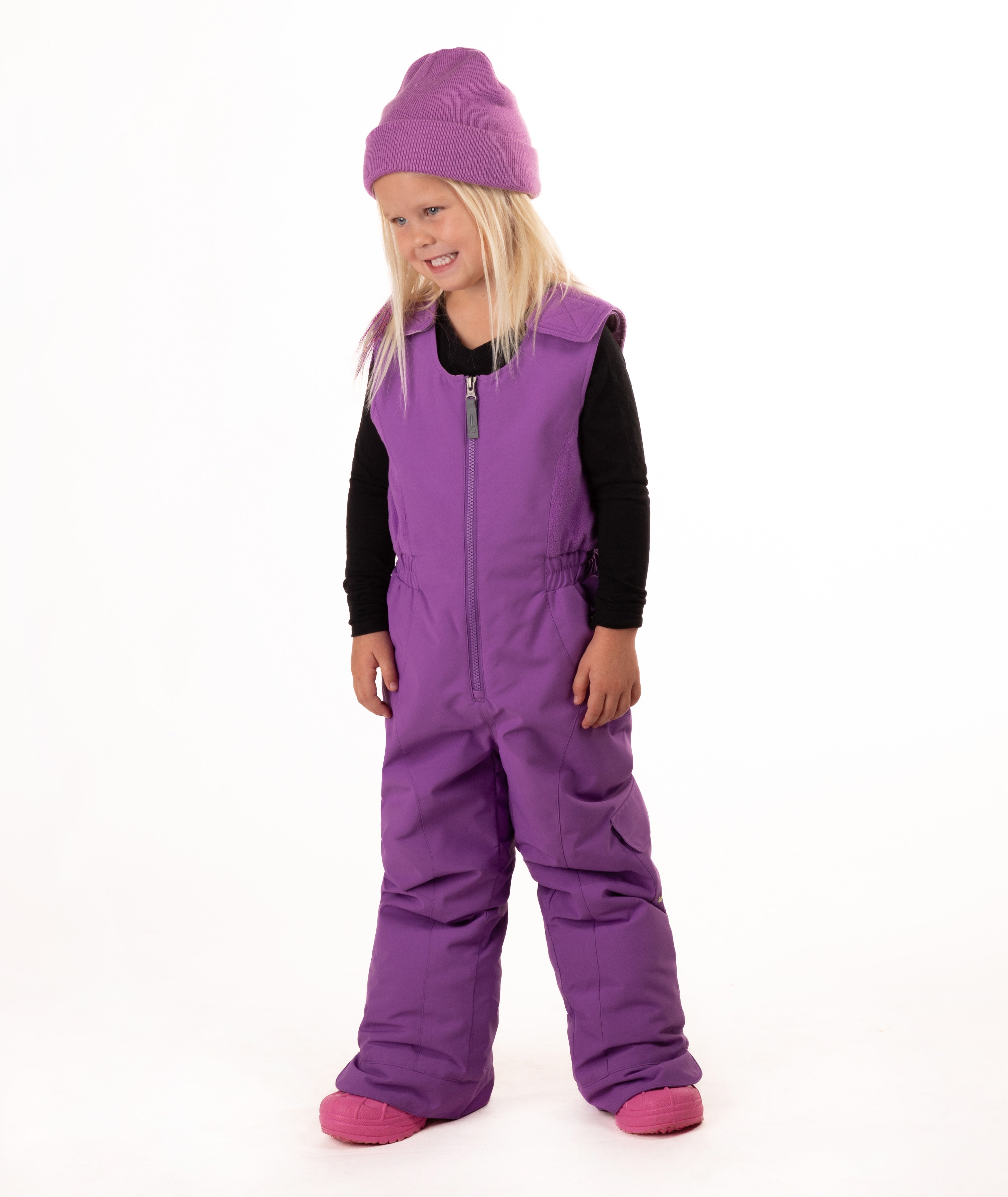 A young child wearing a purple snow bib over a black long-sleeve shirt, a matching purple knit beanie, and pink boots, standing against a plain white studio background.