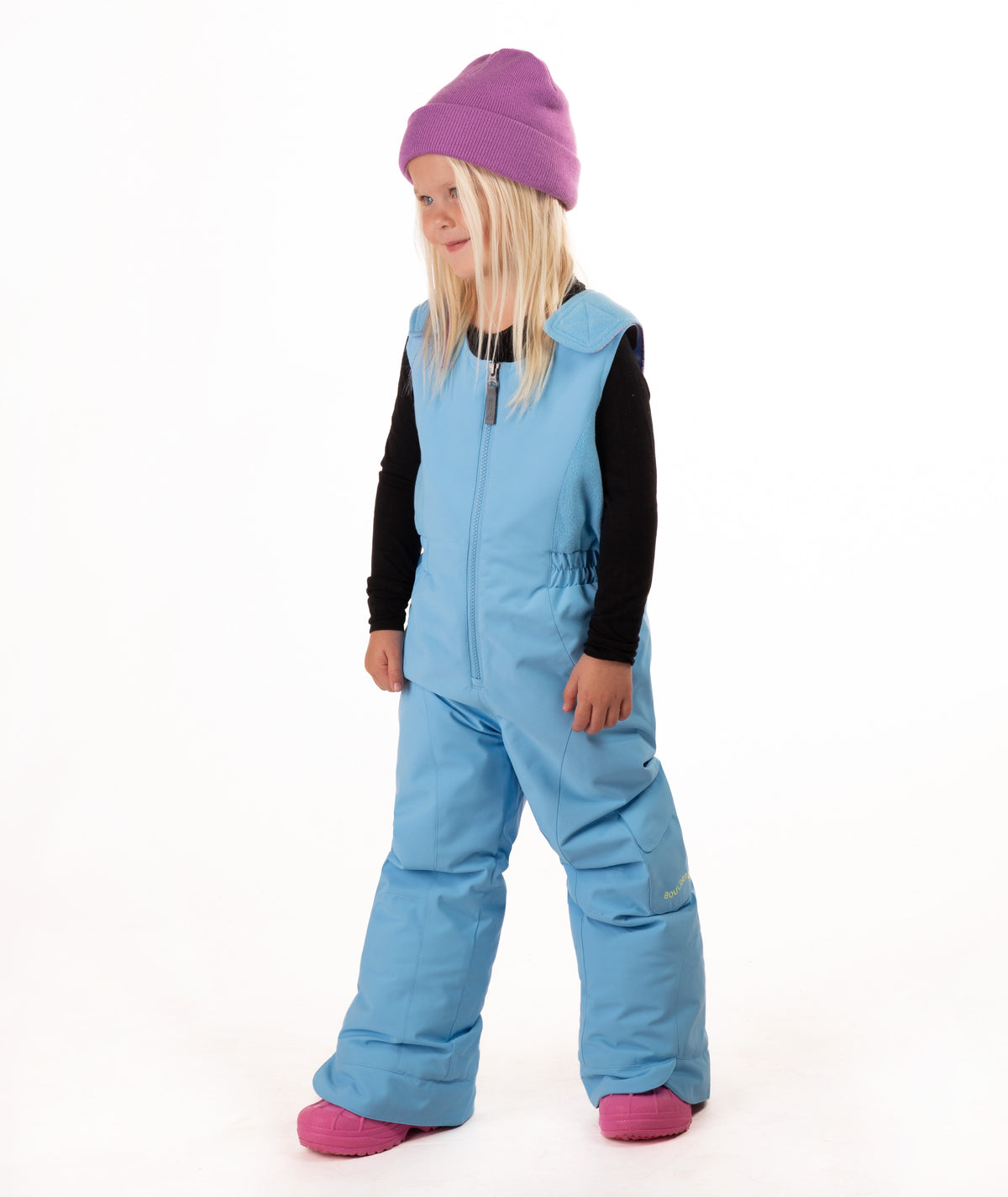 A young child wearing a light blue snow bib over a black long-sleeve shirt, a purple beanie, and pink boots, standing against a plain white studio background.