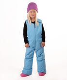 A young child wearing a light blue snow bib over a black long-sleeve shirt, a purple beanie, and pink boots, standing against a plain white studio background.