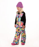 A young child wearing a black long-sleeve top and a colorful abstract print snowsuit with a purple beanie and pink boots, standing against a white studio background.