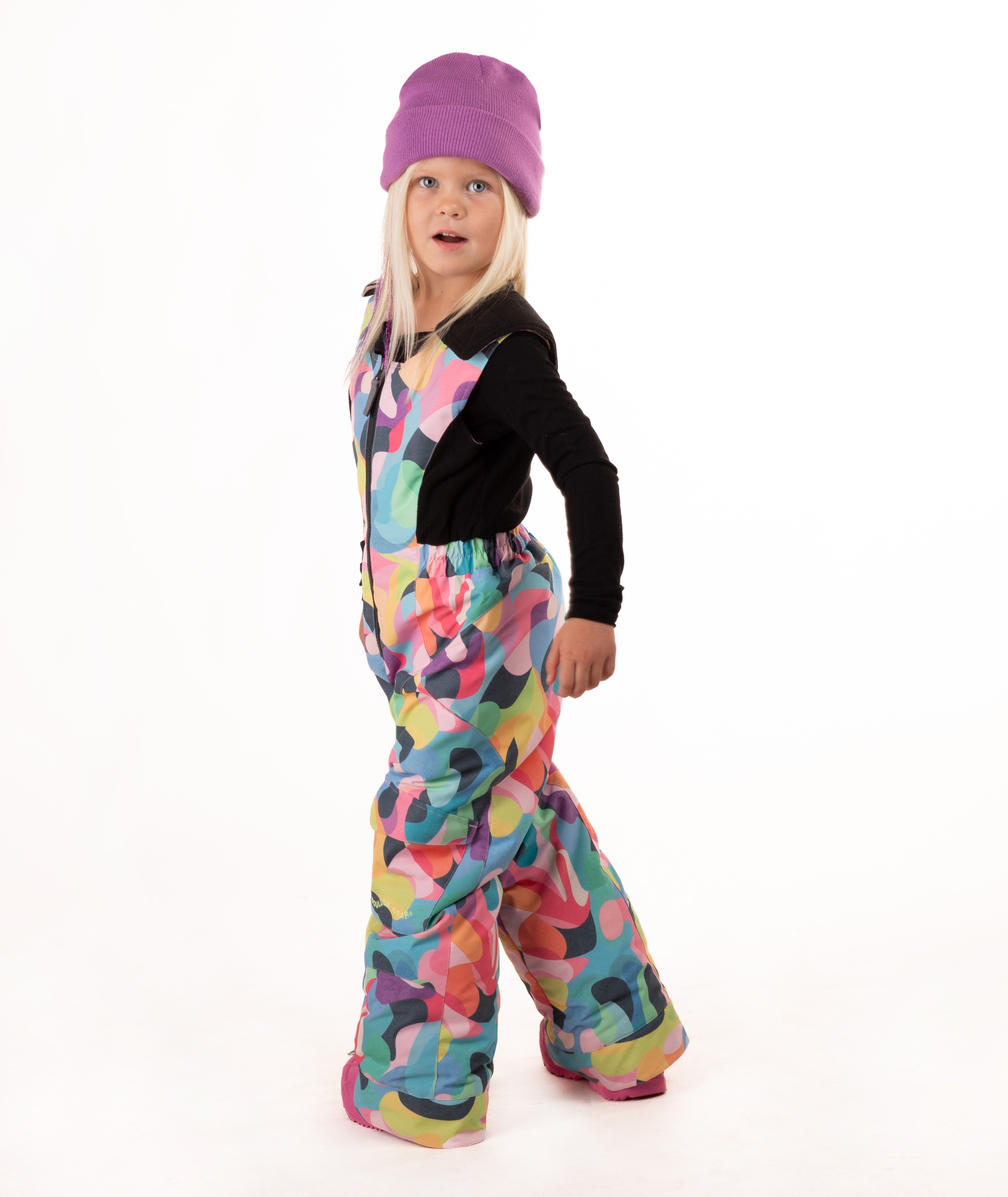 A young child wearing a black long-sleeve top and a colorful abstract print snowsuit with a purple beanie and pink boots, standing against a white studio background.