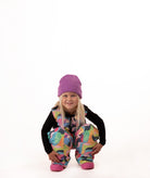 A young girl crouching down in colorful snow pants and a purple beanie, wearing a black long-sleeve shirt, against a solid white background.