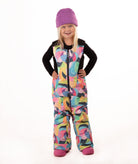 A young child wearing a black long-sleeve top and a colorful abstract print snowsuit with a purple beanie and pink boots, standing against a white studio background.