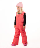 A young girl wearing a pink knit beanie, a black long-sleeve shirt, and bright coral-pink snow bib overalls with matching pink boots, standing against a plain white studio background.
