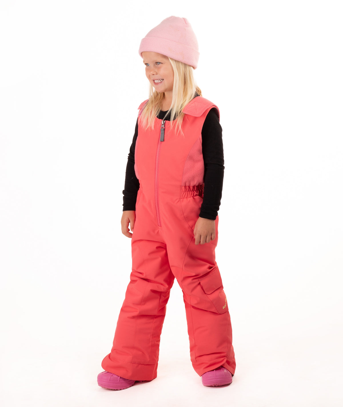 A young girl wearing a pink knit beanie, a black long-sleeve shirt, and bright coral-pink snow bib overalls with matching pink boots, standing against a plain white studio background.
