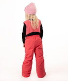 A young girl wearing a pink knit beanie, a black long-sleeve shirt, and bright coral-pink snow bib overalls with matching pink boots, standing against a plain white studio background.