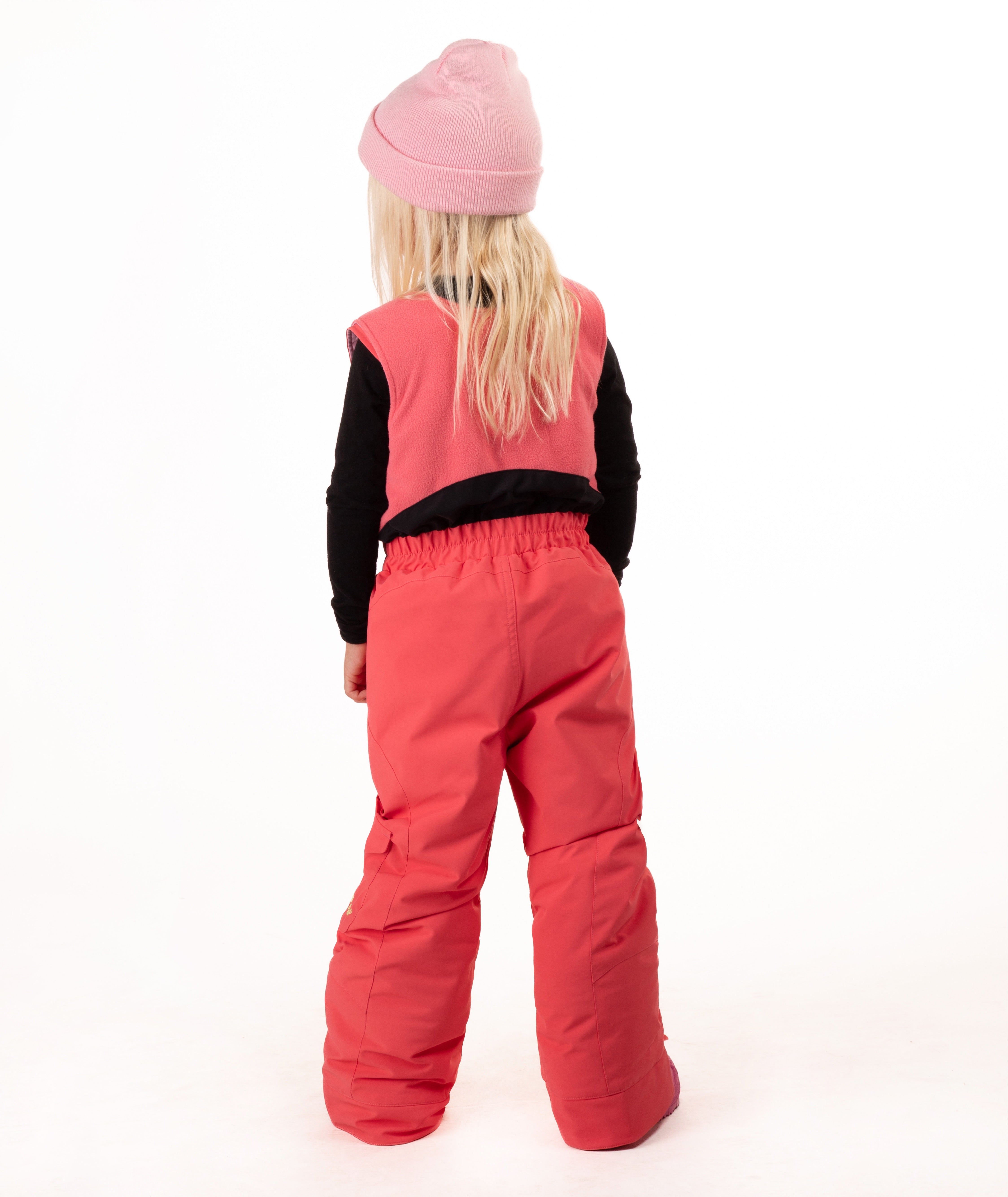 A young girl wearing a pink knit beanie, a black long-sleeve shirt, and bright coral-pink snow bib overalls with matching pink boots, standing against a plain white studio background.