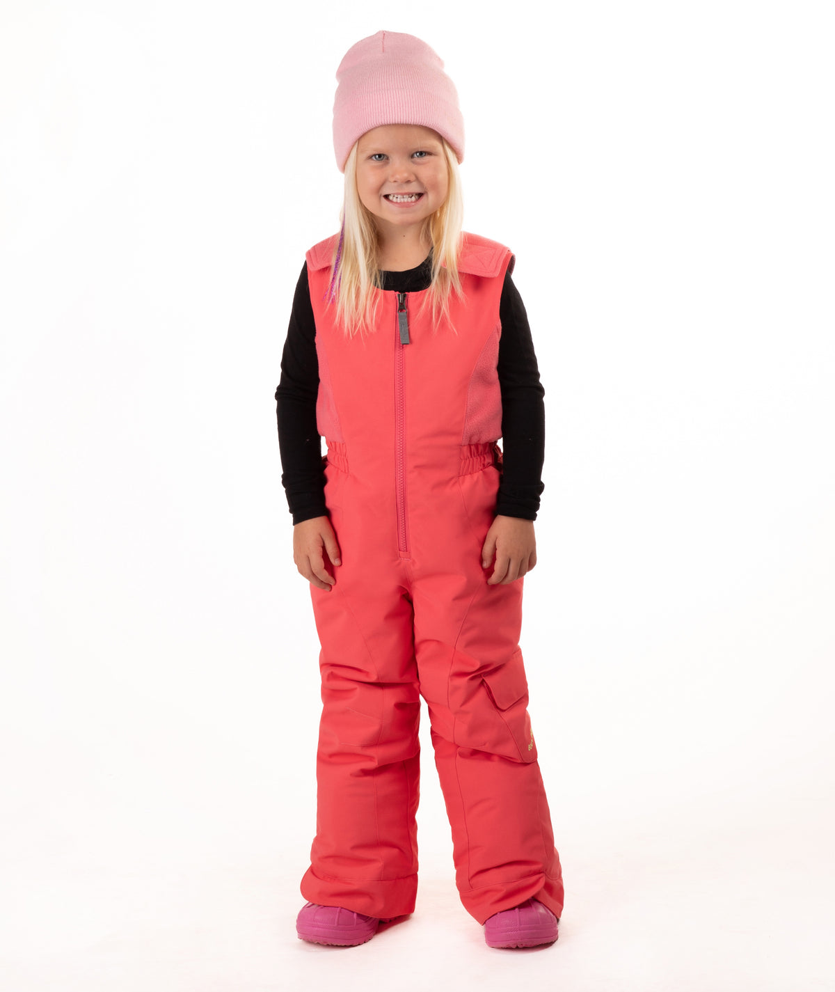 A young girl wearing a pink knit beanie, a black long-sleeve shirt, and bright coral-pink snow bib overalls with matching pink boots, standing against a plain white studio background.