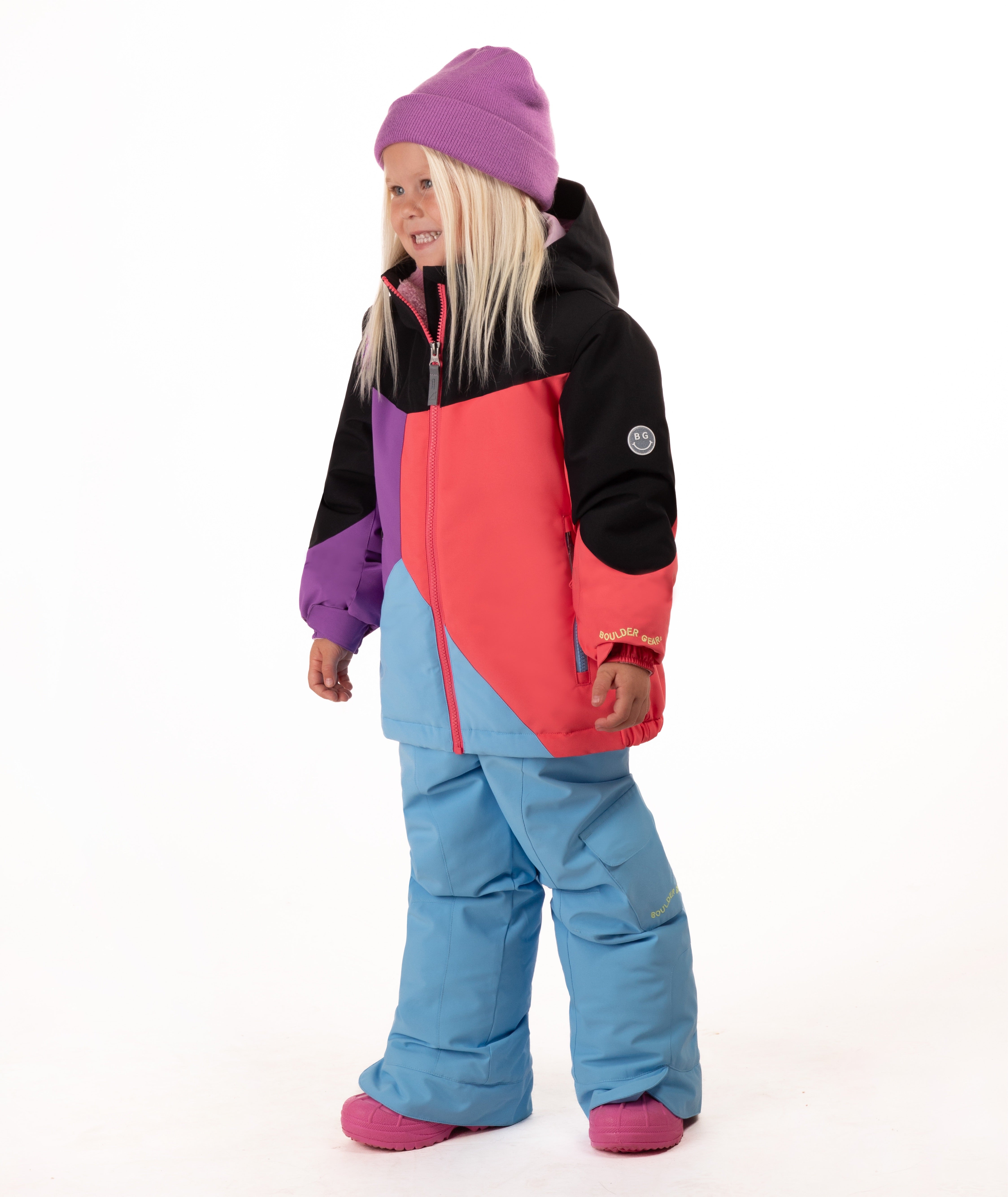 A young child wearing a colorful color-blocked ski jacket, light blue snow pants, a purple beanie, and pink boots, standing against a plain white studio background.