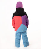 A young child wearing a colorful color-blocked ski jacket, light blue snow pants, a purple beanie, and pink boots, standing against a plain white studio background.