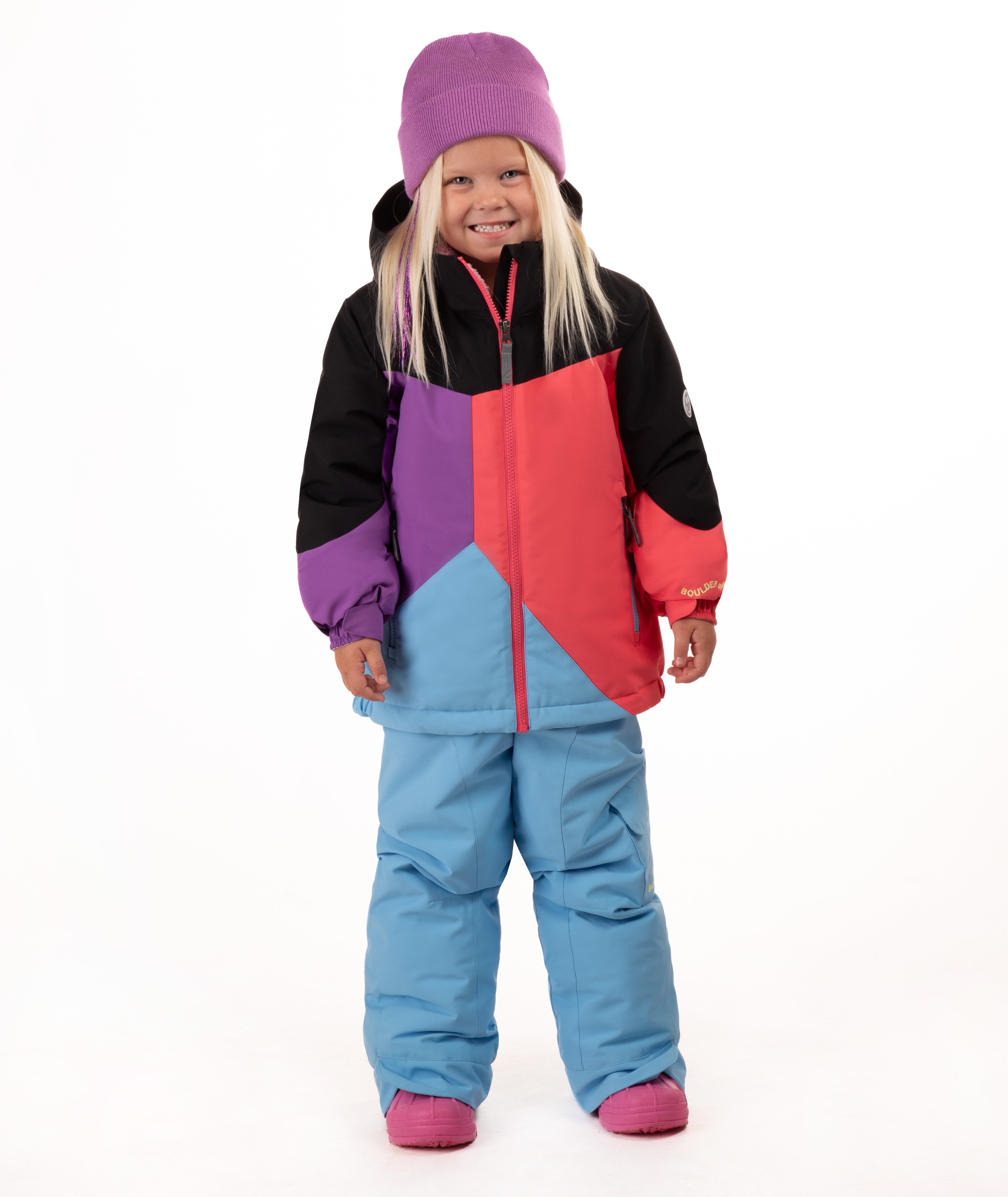 A young child wearing a colorful color-blocked ski jacket, light blue snow pants, a purple beanie, and pink boots, standing against a plain white studio background.