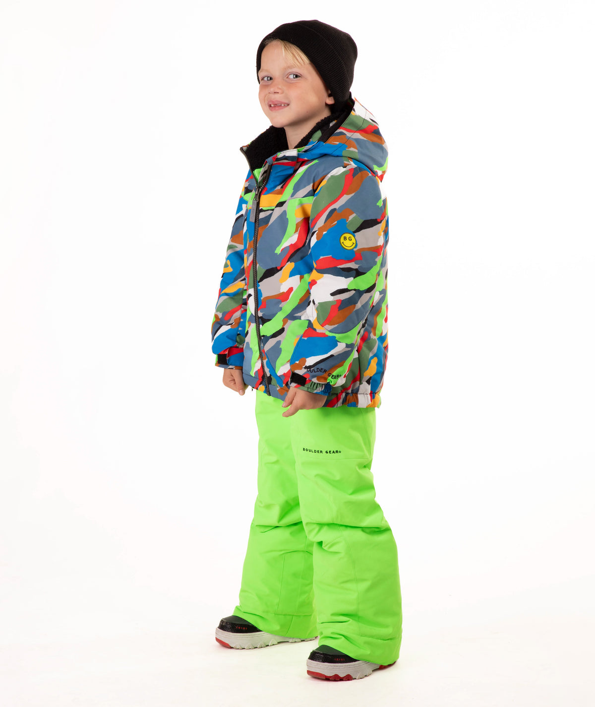 A child wearing a colorful camouflage-patterned ski jacket and bright neon green snow pants, standing against a plain white studio background.