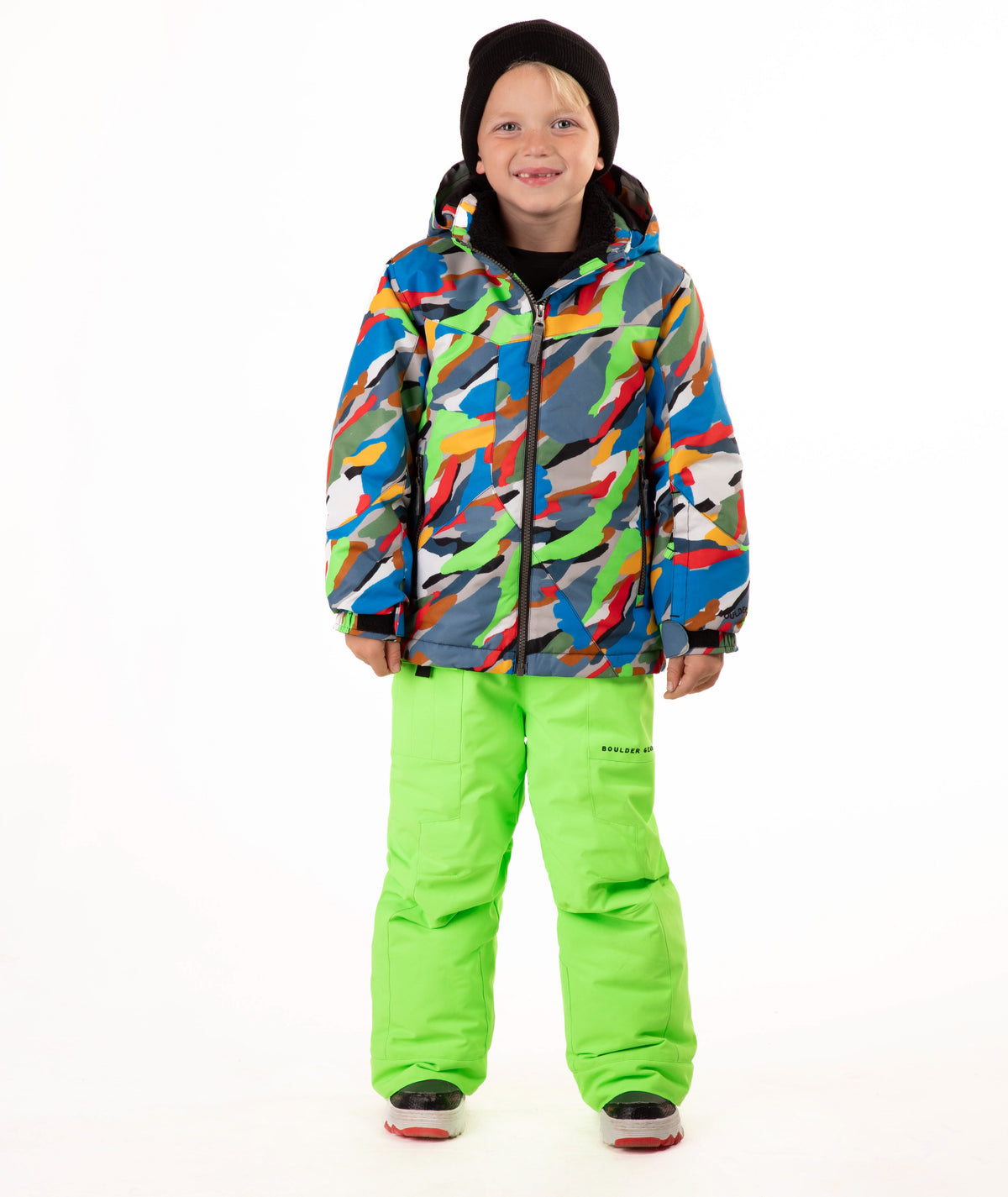 A child wearing a colorful camouflage-patterned ski jacket and bright neon green snow pants, standing against a plain white studio background.